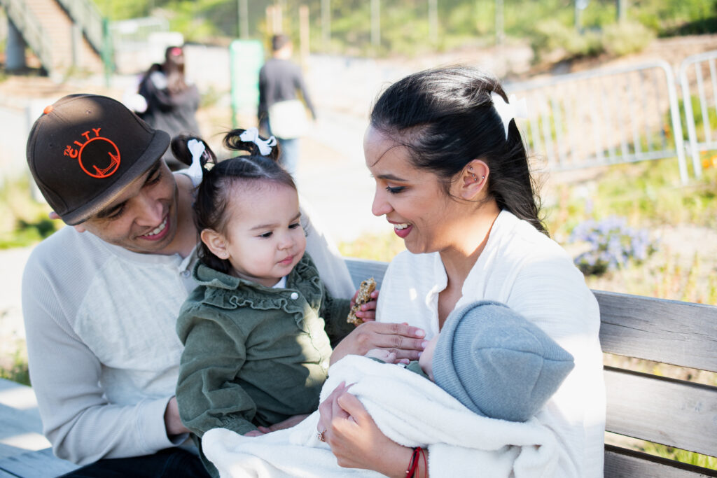 mother holding a baby with father and toddle child sitting on park bench