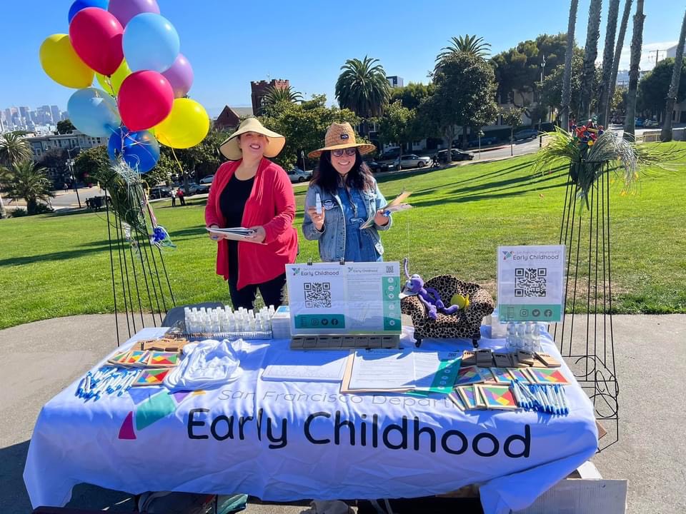 two women standing with balloons at a table with a DEC tablecloth on it in a park